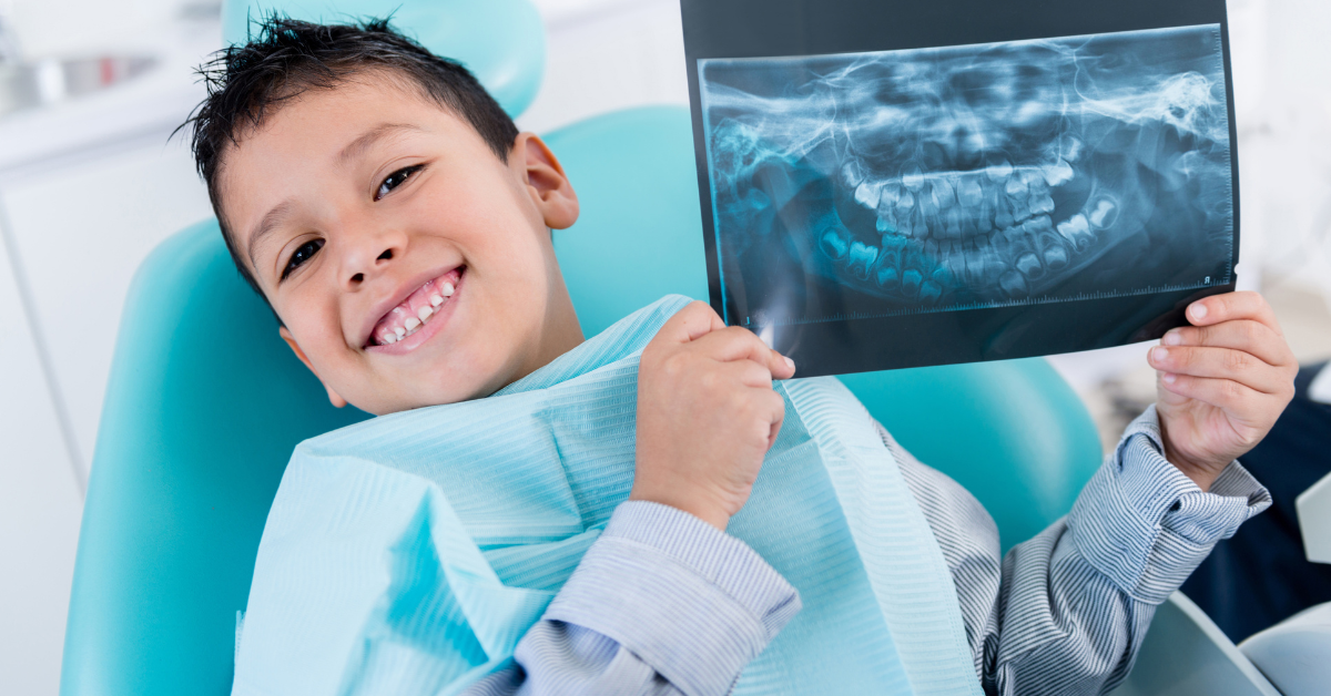 A smiling young boy sits in a dentist's chair, proudly holding up a dental X-ray. The bright setting conveys a cheerful and reassuring tone.