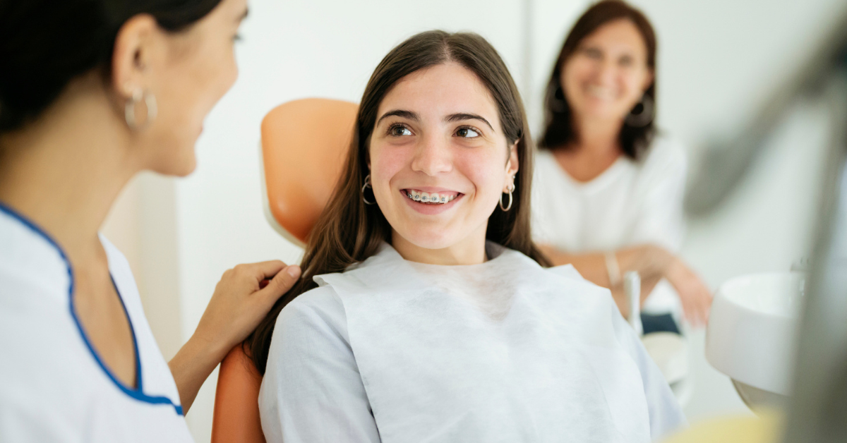 Teen girl with braces in a dentist's chair smiles at a female dentist. Another woman sits in the background. The mood is friendly and reassuring.