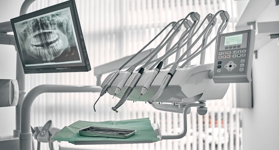 A dental office setup featuring a screen displaying a dental X-ray of teeth, dental tools on a tray with a green cover, and an equipment panel, in a bright room.