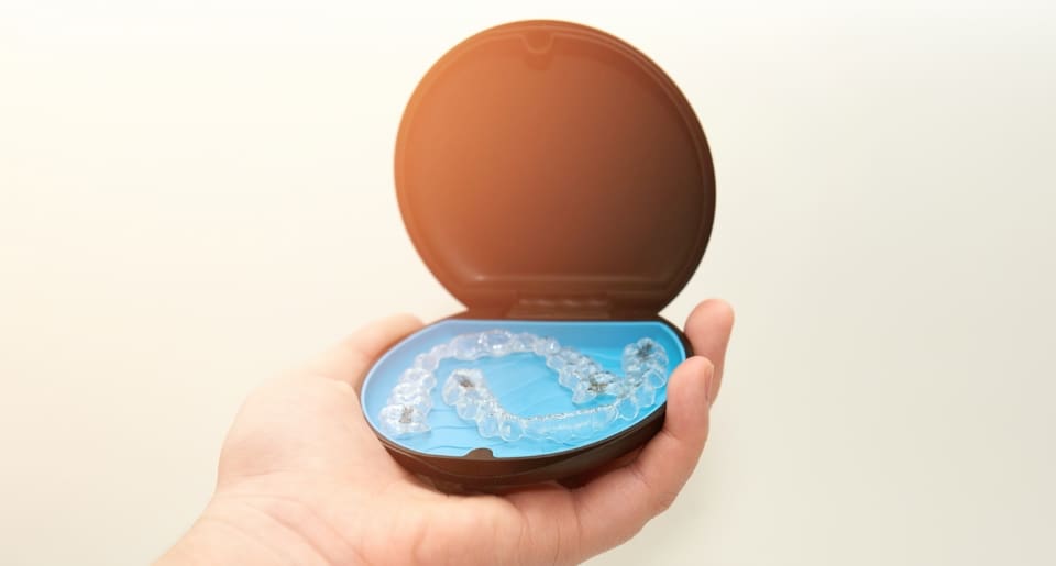 A hand holds an open dark case containing clear dental aligners on a blue interior. The image is well-lit, giving a clean, clinical feel.