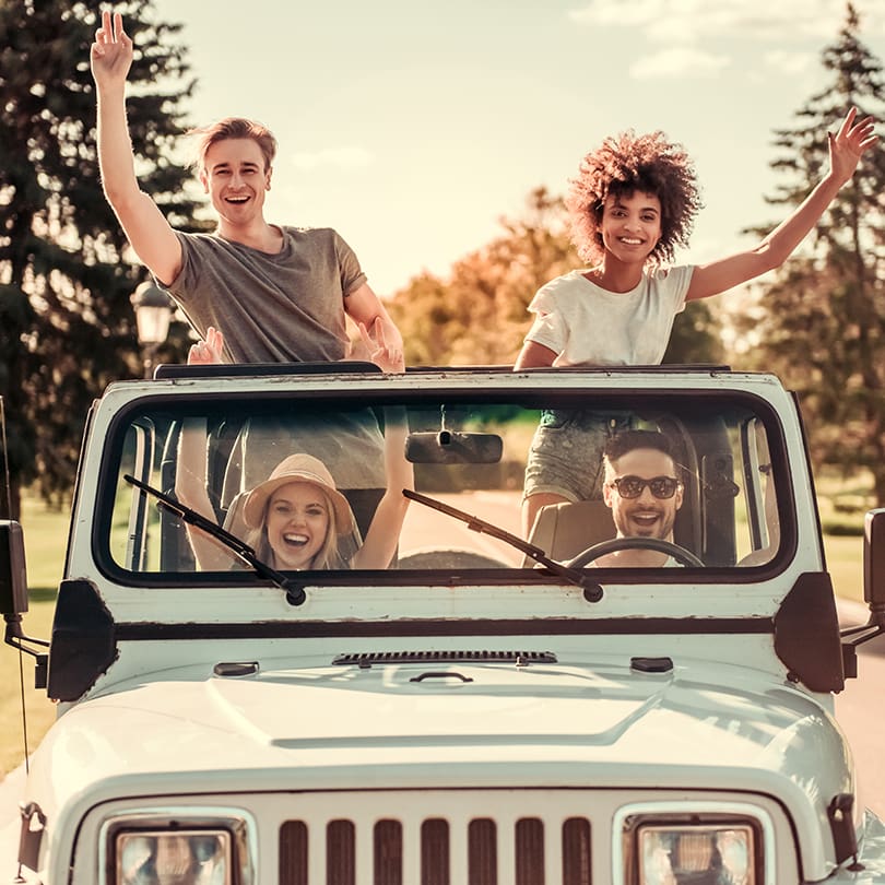 A group of four friends enjoying a sunny day, standing and sitting in a white convertible Jeep. They are smiling, with hands raised joyfully. Trees in the background.