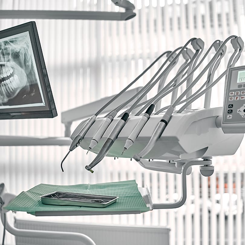 A dentist's office with a display showing a dental X-ray and a dental tool tray. The image conveys a clean, clinical atmosphere.