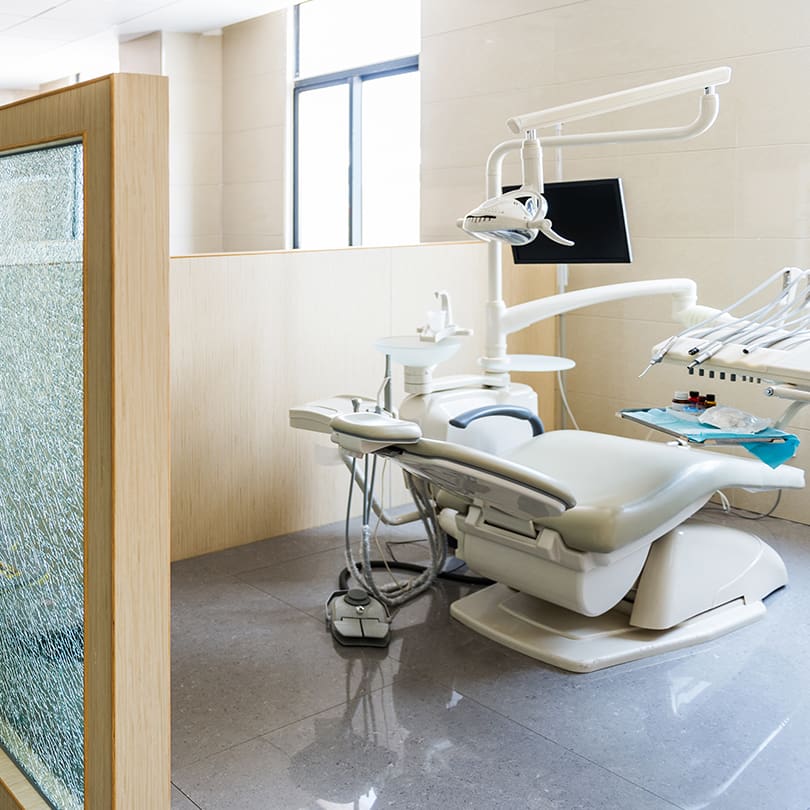 Dental office with a modern chair, overhead light, and monitor. The room is bright, with a frosted glass partition and beige walls, creating a clean, welcoming atmosphere.