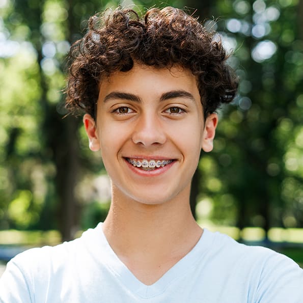 Smiling teenager with curly hair and braces, wearing a light blue shirt. He stands in a sunny park with trees blurred in the background, conveying a cheerful mood.